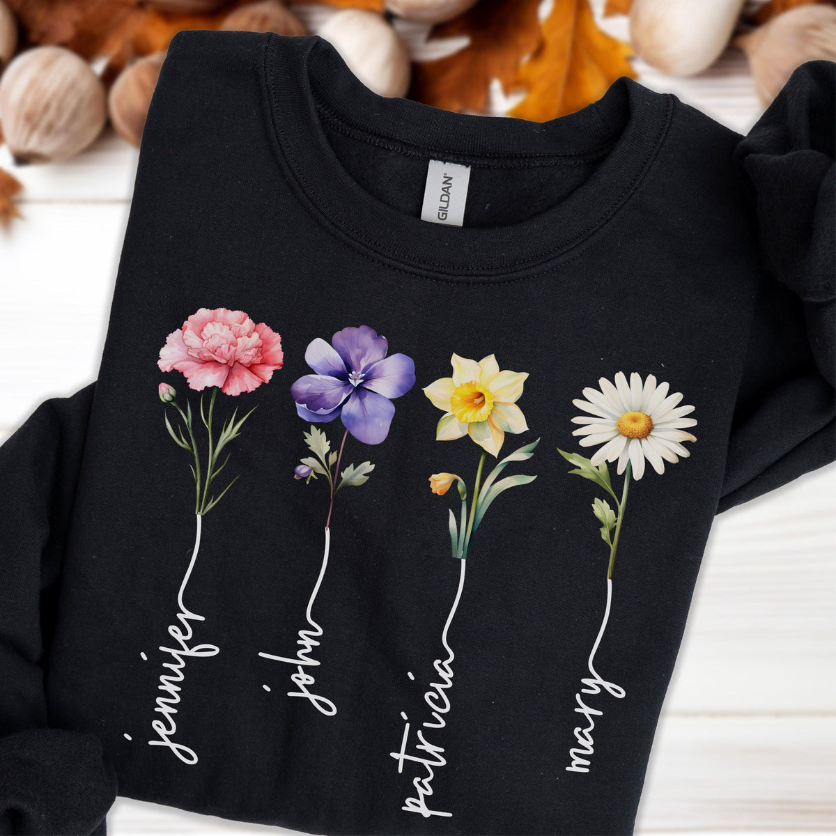 May include: Black sweatshirt featuring four colorful floral illustrations: a pink carnation, a purple pansy, a yellow daffodil, and a white daisy. Each flower has a name written below it in a cursive font: Jennifer, John, Patricia, and Mary.