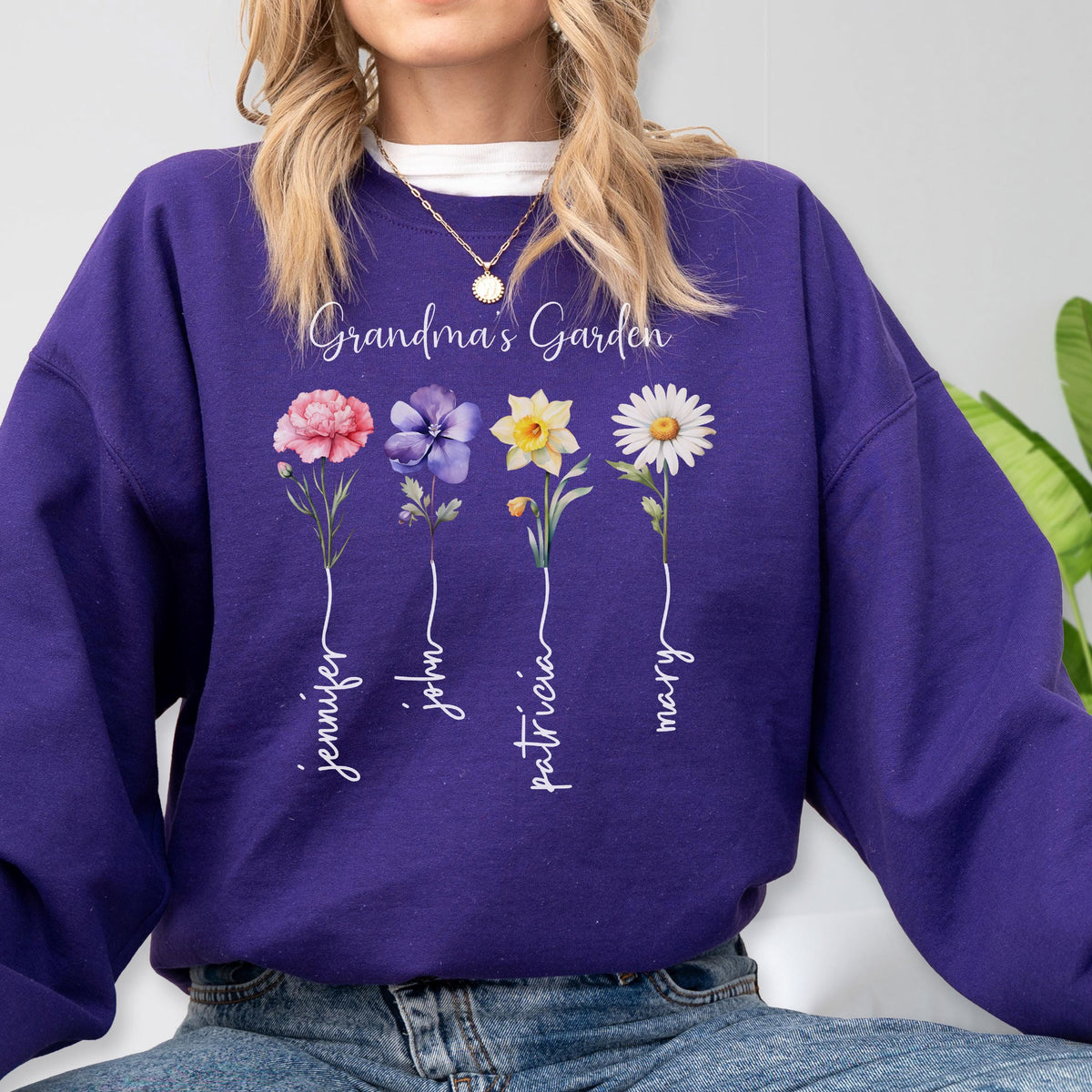 May include: A purple sweatshirt featuring the text "Grandma's Garden" above four watercolor flowers: a pink rose, a purple flower, a yellow daffodil, and a white daisy. Each flower has a name written below it in cursive.
