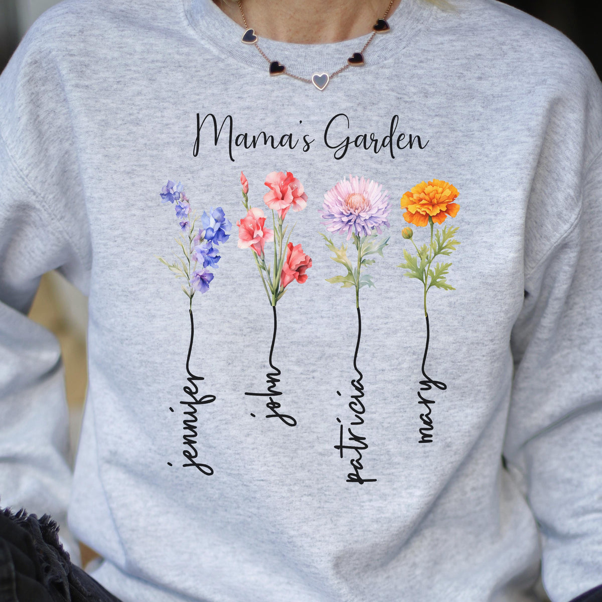 May include: A light gray sweatshirt with the text "Mama's Garden" and four colorful flower illustrations. Each flower has a name written below it: Jennifer, John, Patricia, and Mary. The flowers are blue, red, pink, and orange.