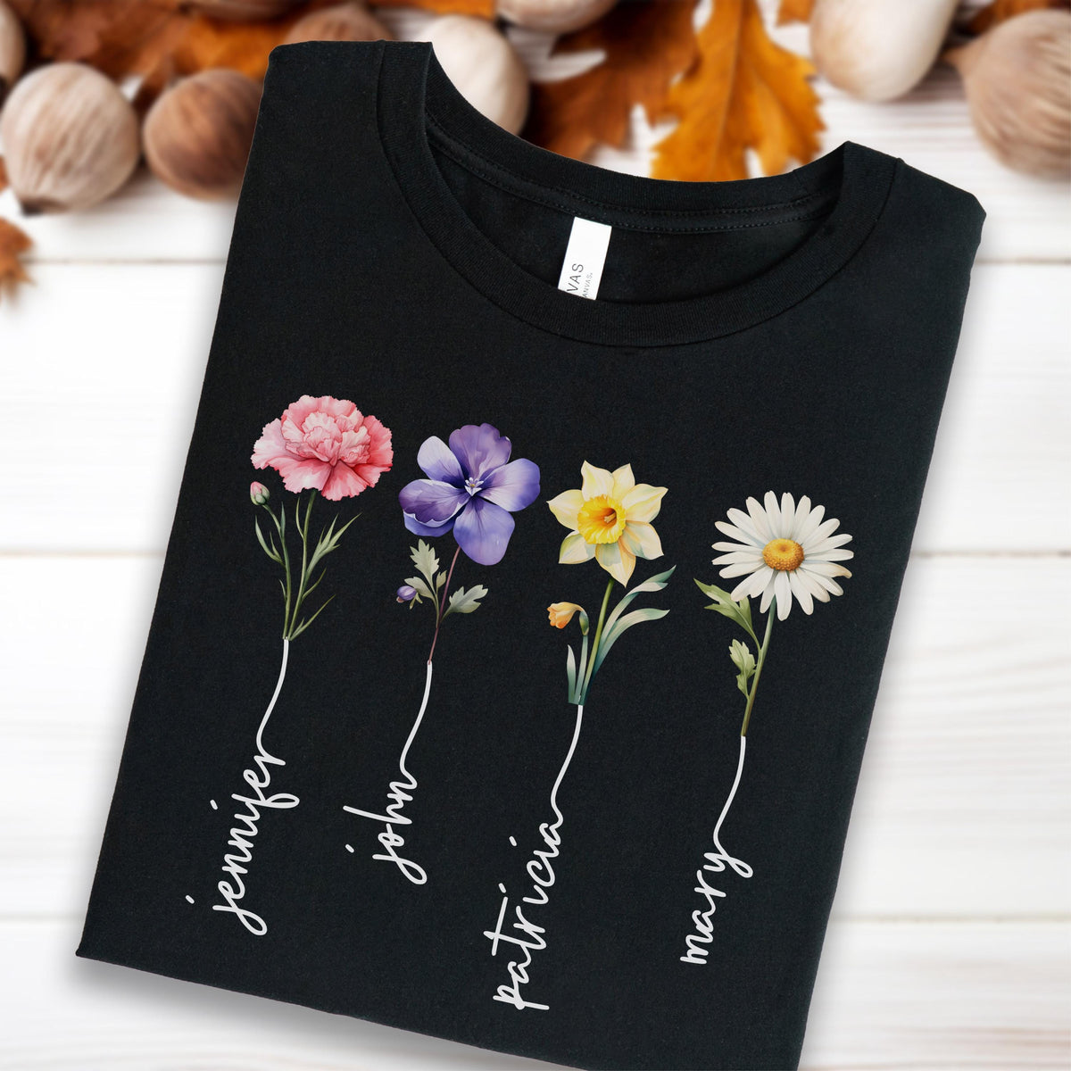 May include: Black t-shirt featuring watercolor flowers in pink, purple, yellow, and white. The names "Jennifer", "John", "Patricia", and "Mary" are written in white script below each flower. The shirt is made of a soft material.