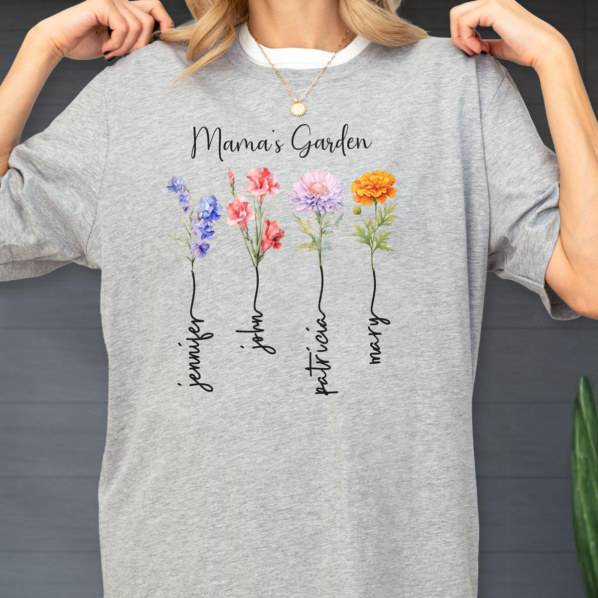 May include: A light gray t-shirt featuring the text "Mama's Garden" above four watercolor flowers. Each flower has a name written below it: Jennifer, John, Patricia, and Mary. The flowers are blue, pink, purple, and orange.