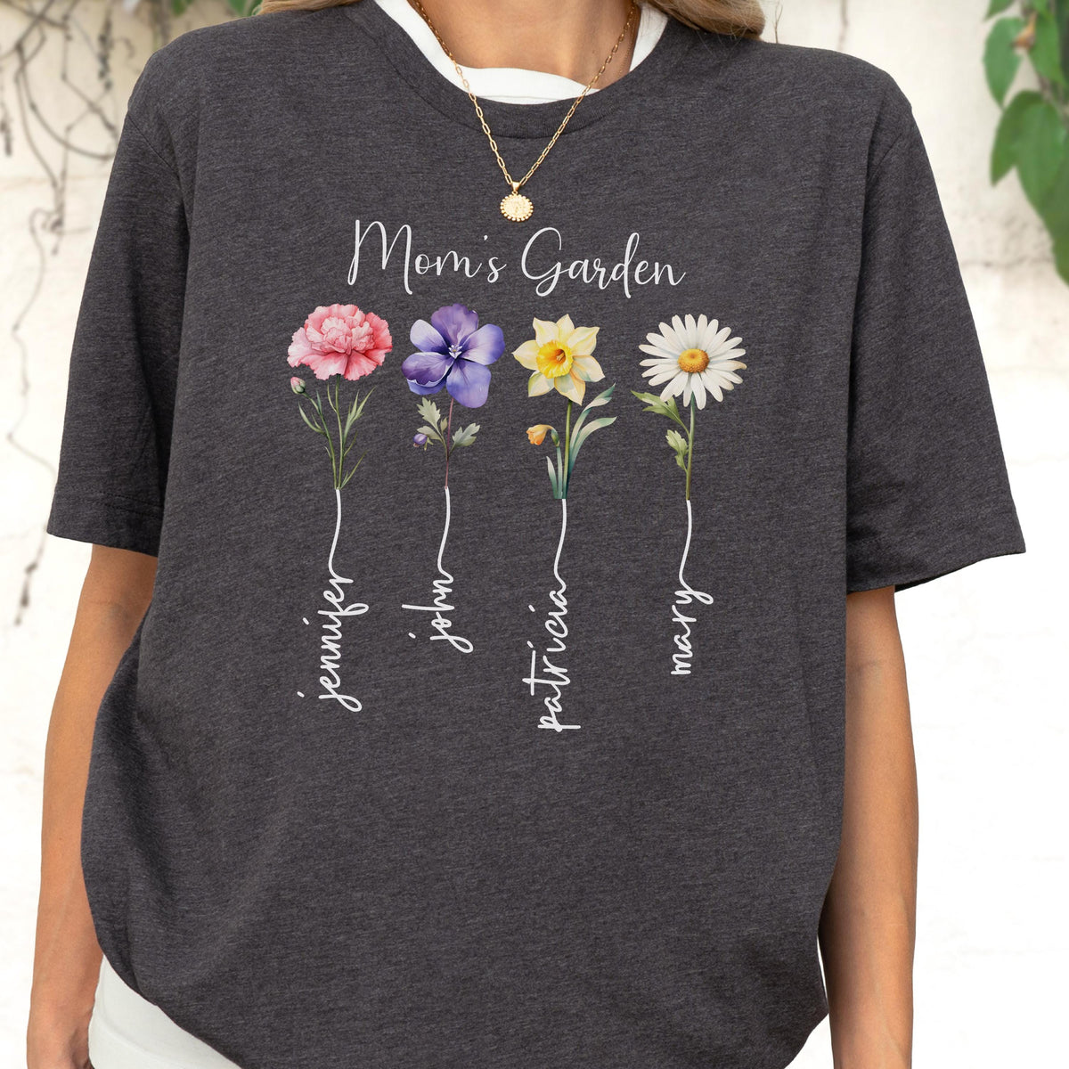 May include: Dark gray t-shirt with the text "Mom's Garden" and four watercolor flowers: a pink peony, a purple flower, a yellow daffodil, and a white daisy. Names Jennifer, John, Patricia, and Mary are written below each flower.
