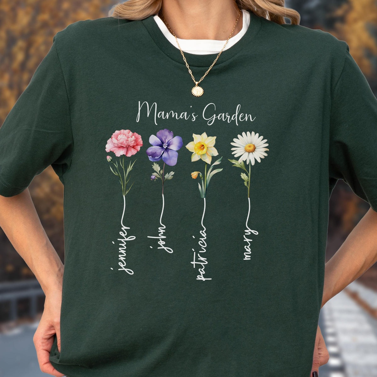 May include: Dark green t-shirt with the text "Mama's Garden" and four flower illustrations. Each flower has a name written below it: Jennifer, John, Patricia, and Mary. The shirt has a white crew neck.