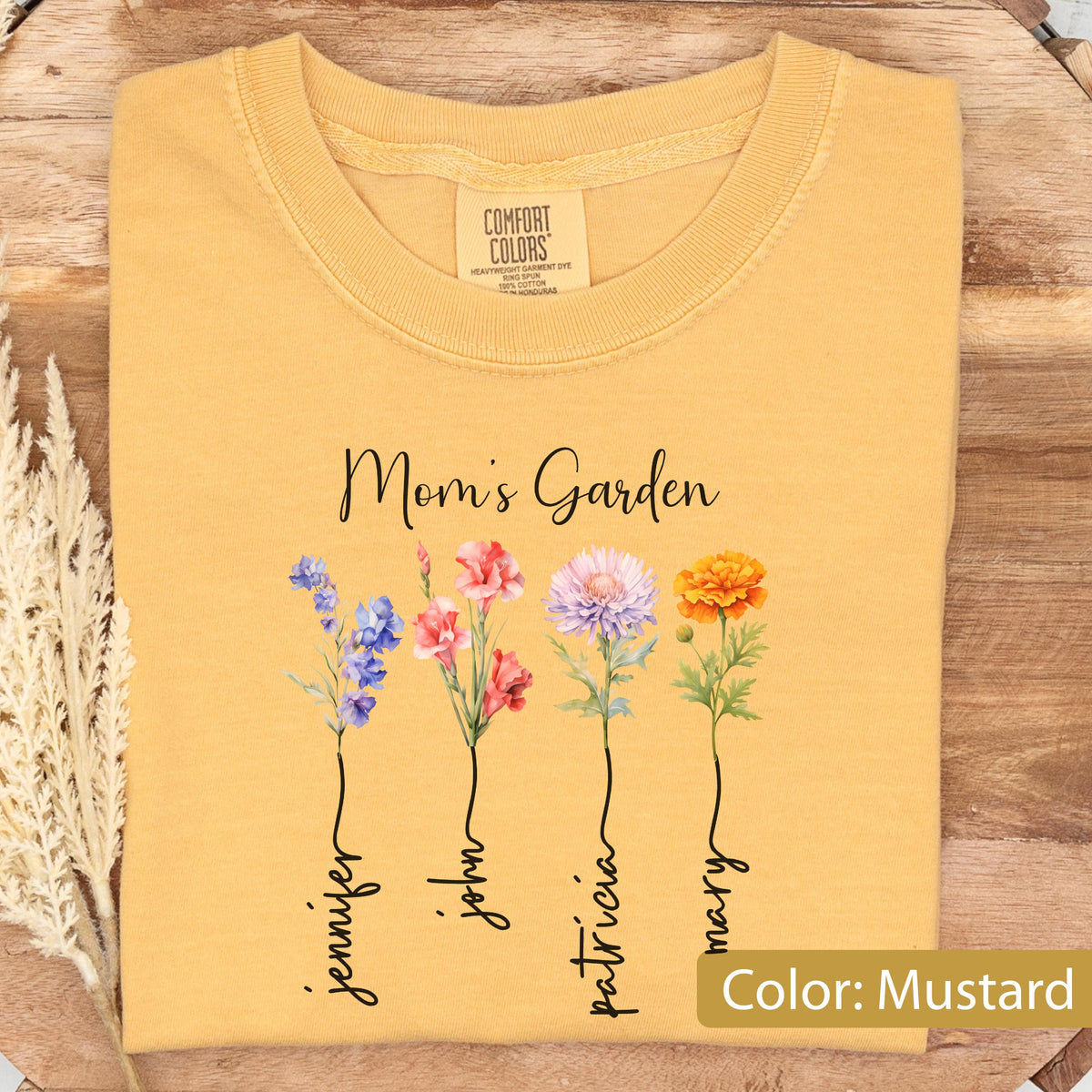 May include: Mustard-colored t-shirt with the text "Mom's Garden" and floral illustrations. The shirt features four flowers with names: Jennifer, John, Patricia, and Mary. The shirt is made of a soft material.