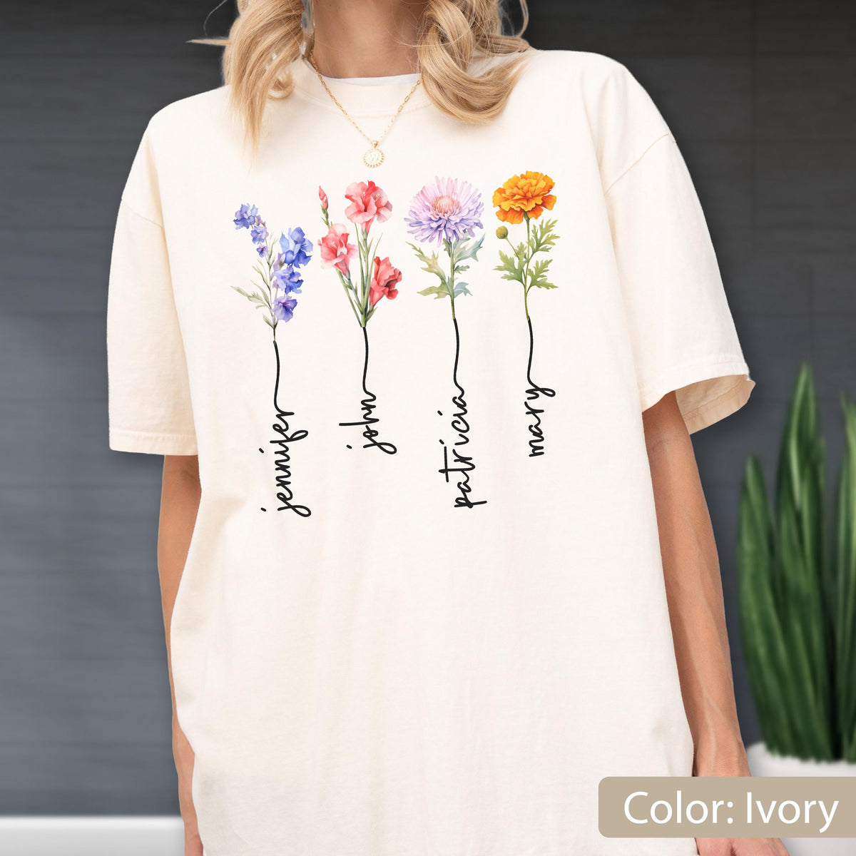 May include: Ivory-colored t-shirt featuring watercolor floral designs with the names "Jennifer", "John", "Patricia", and "Mary" in a script font. The flowers are in shades of blue, red, pink, purple, and orange. The shirt is a relaxed fit.