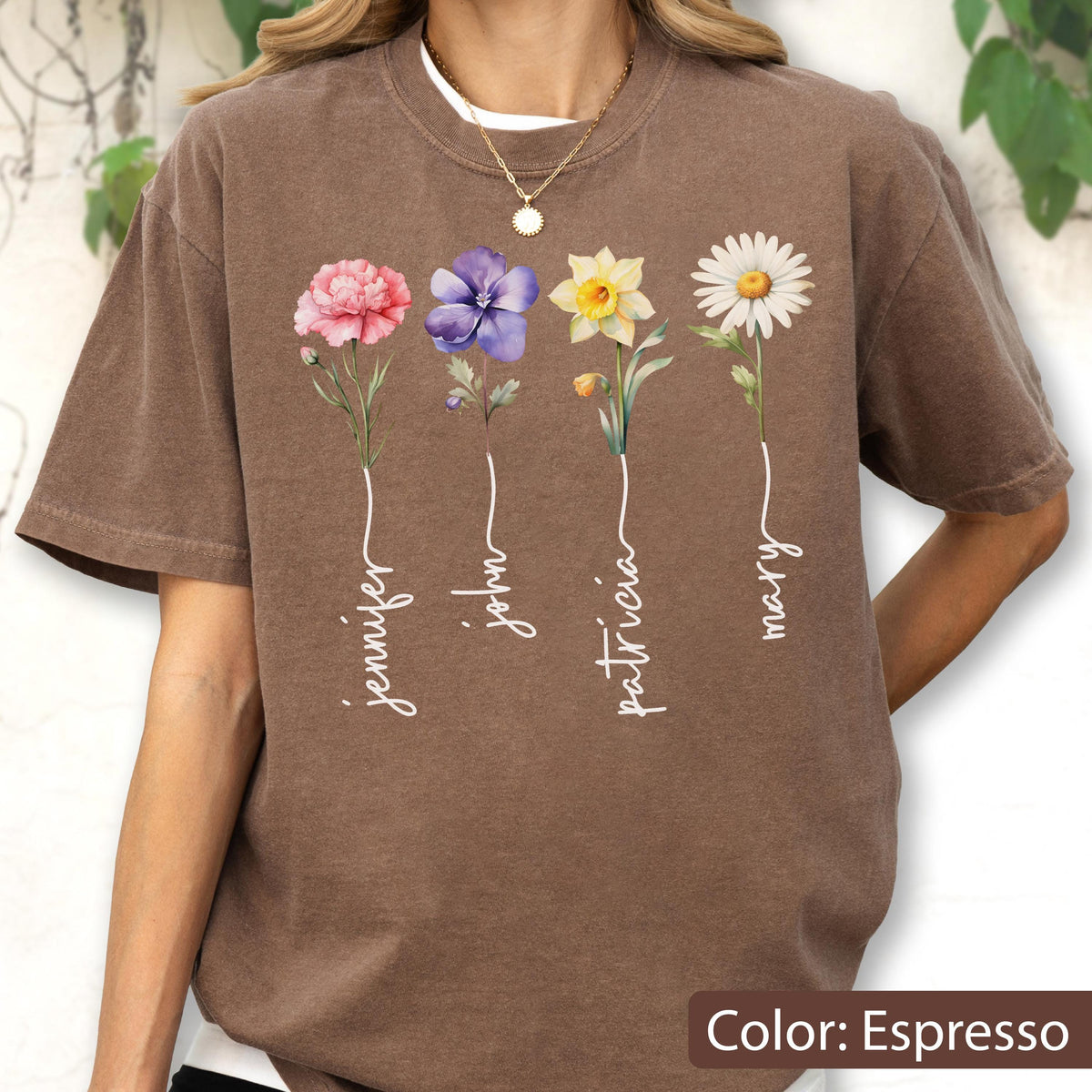 May include: Espresso-colored t-shirt featuring four floral designs with names: Jennifer, John, Patricia, and Mary. Each flower is a different color: pink, purple, yellow, and white. The shirt has a crew neck and short sleeves.