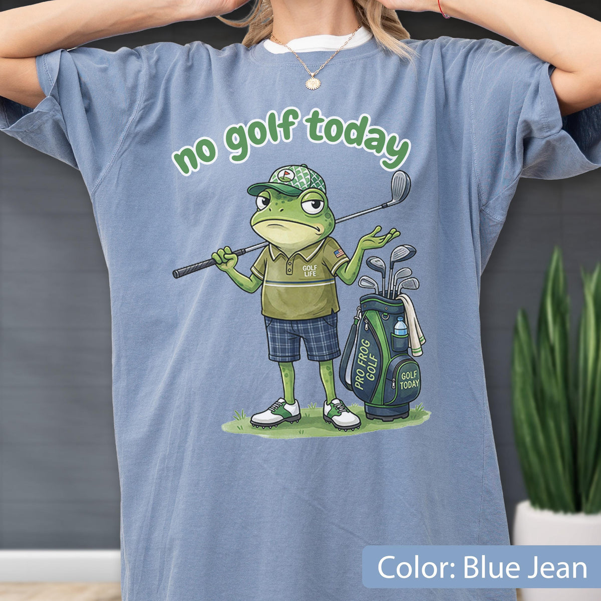 May include: Blue jean colored t-shirt featuring a cartoon frog golfer with the text "no golf today". The frog is wearing a golf hat, polo shirt, and plaid shorts, holding a golf club. A golf bag with clubs is next to the frog.