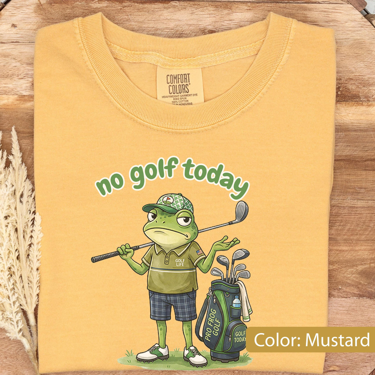 May include: Mustard-colored t-shirt featuring a cartoon frog golfer with the text "no golf today." The frog is wearing a golf cap, polo shirt, and plaid shorts, holding a golf club. A golf bag with clubs is next to the frog.