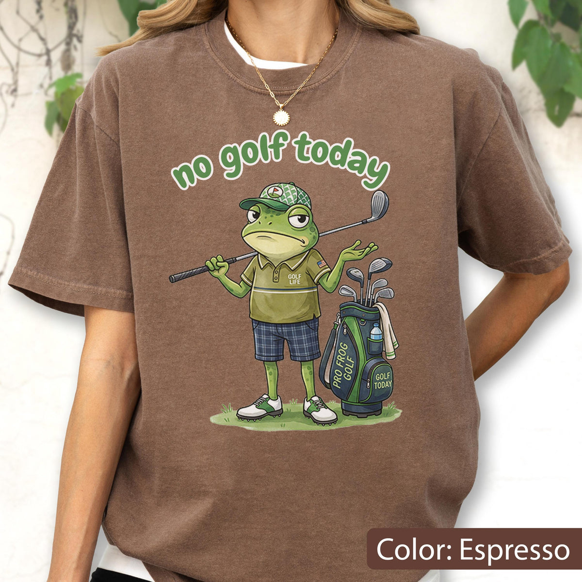 May include: Espresso-colored t-shirt featuring a cartoon frog golfer with the text "no golf today." The frog wears a golf cap, polo shirt, and shorts, holding a golf club. A golf bag with clubs is beside him.