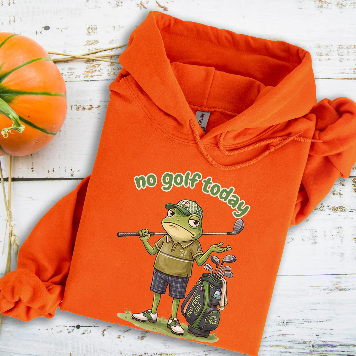 May include: Orange hoodie with the text "no golf today" in green. The hoodie features a cartoon frog wearing a golf hat and holding a golf club. A golf bag with clubs is next to the frog. The hoodie is on a light wood background.
