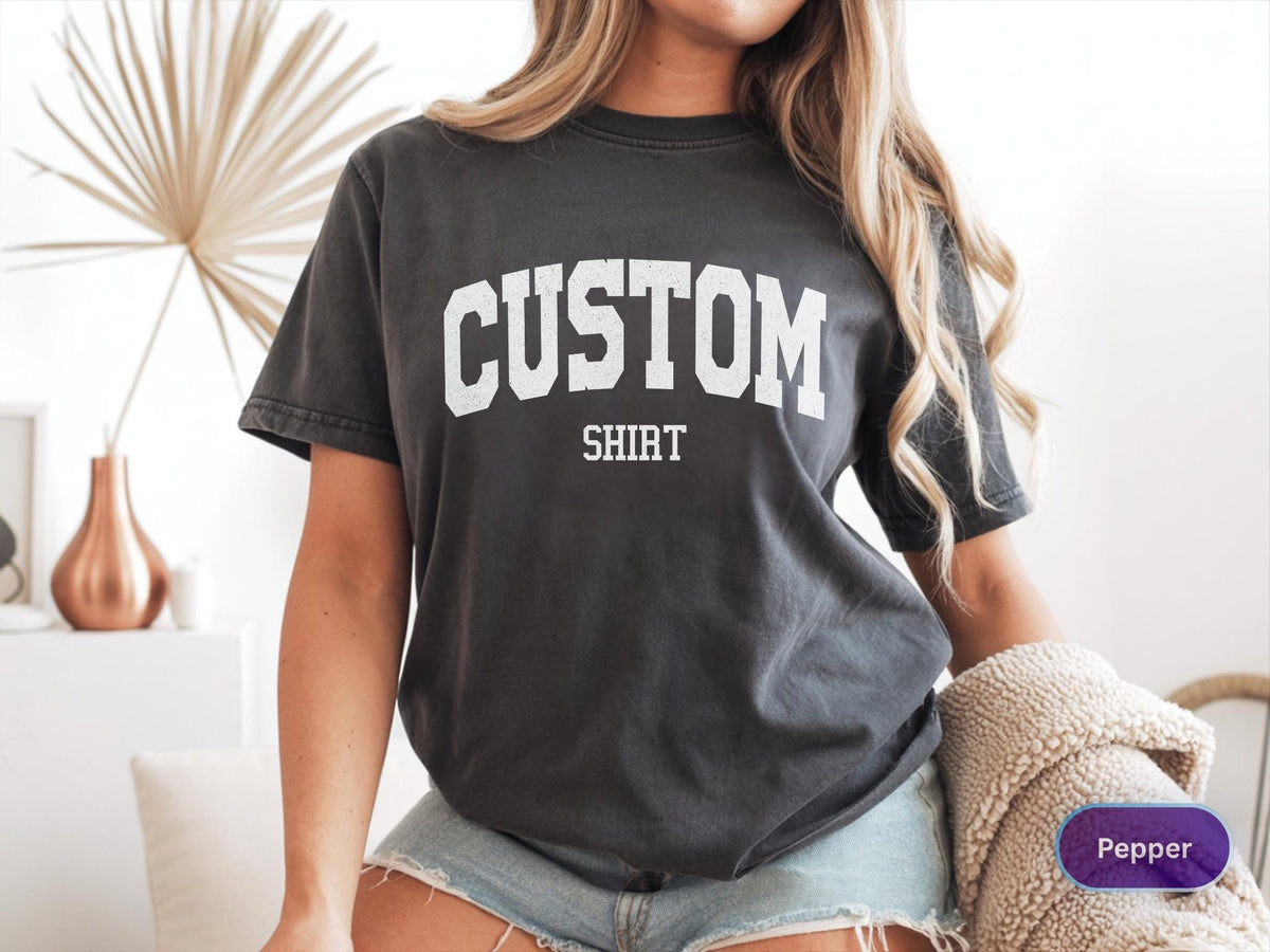 May include: Dark gray t-shirt with the words "CUSTOM SHIRT" in white block letters. The shirt is a crew neck style and appears to be made of a soft, comfortable material. The shirt is worn with denim shorts.