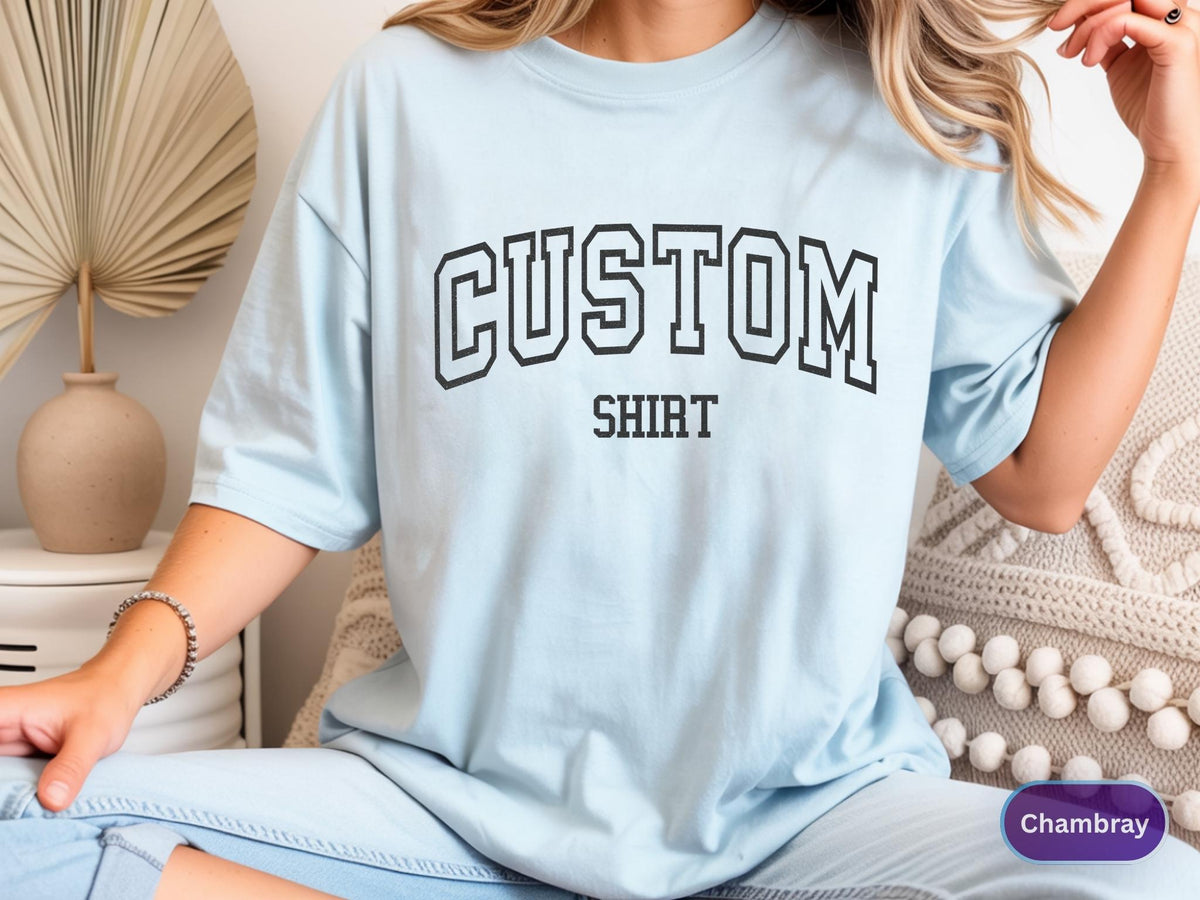 May include: Light blue t-shirt with the word "CUSTOM" above "SHIRT" in black block letters. The shirt is a soft, casual style.