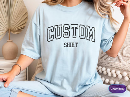 May include: Light blue t-shirt with the word "CUSTOM" above "SHIRT" in black block letters. The shirt is a soft, casual style.