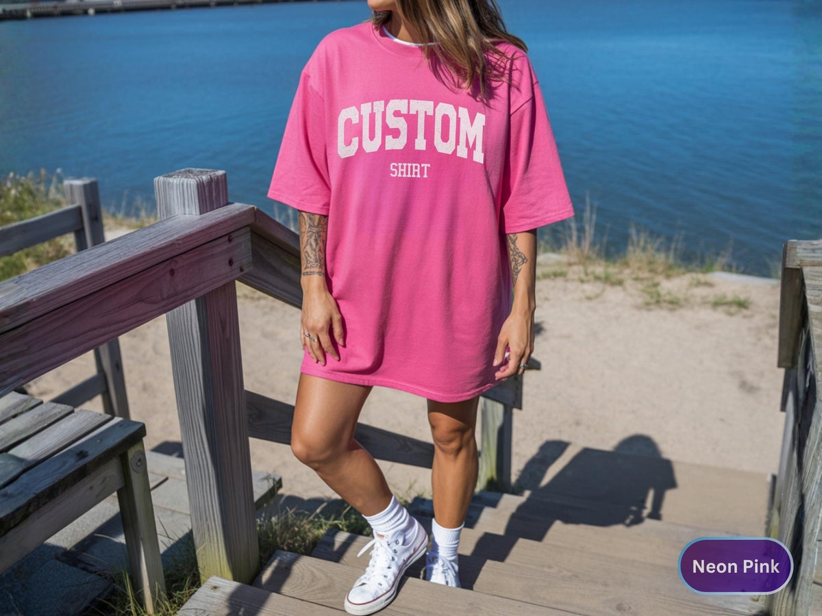 May include: A neon pink oversized t-shirt with the word "CUSTOM" printed in white block letters. The word "SHIRT" is printed below "CUSTOM". The shirt is worn with white sneakers and socks. The background includes a wooden staircase and a body of water.