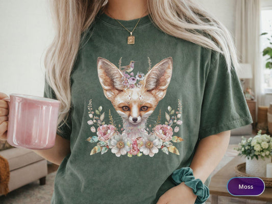 May include: Moss green t-shirt featuring a watercolor illustration of a fox with large ears, adorned with flowers and a small bird. The fox has a white face and brown fur. The design is surrounded by pink and blue flowers.