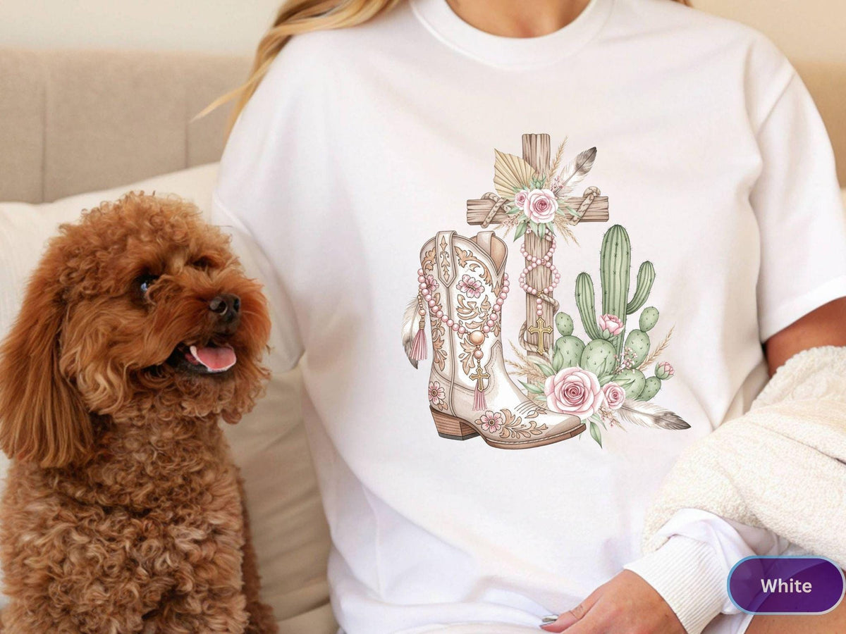 May include: White t-shirt featuring a watercolor design of a cross, cowboy boot, and cactus with pink roses and feathers. The design has a rustic, country theme. A small, brown dog is in the image.