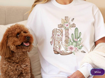 May include: White t-shirt featuring a watercolor design of a cross, cowboy boot, and cactus with pink roses and feathers. The design has a rustic, country theme. A small, brown dog is in the image.
