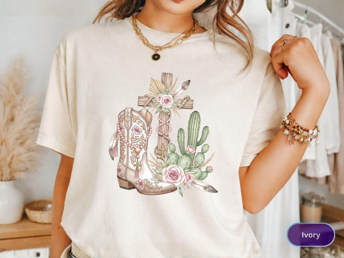 May include: Ivory-colored t-shirt featuring a watercolor design of a wooden cross, a cowboy boot, and a cactus. The design includes pink roses and decorative elements. The shirt is a casual, comfortable style.