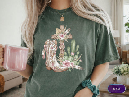May include: Moss green t-shirt featuring a detailed graphic of a cowboy boot, cross, cactus, and floral elements. The boot is cream-colored with intricate designs. The cross is wooden, and the design includes pink roses and feathers.