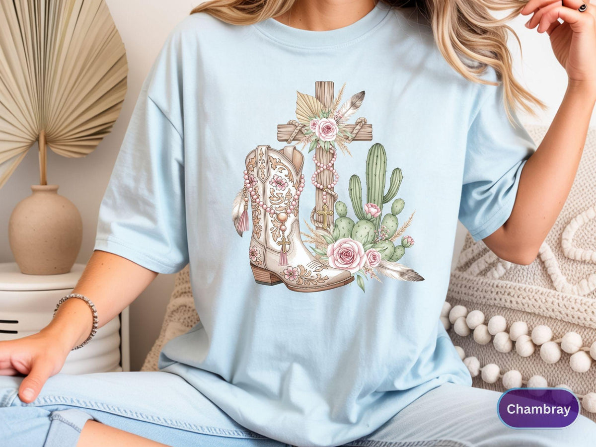 May include: Light blue t-shirt featuring a watercolor design of a cross, cowboy boot, and cactus. The boot is white and brown with floral details. The cross is made of wood and adorned with flowers and feathers. The word "Chambray" is in the bottom right corner.
