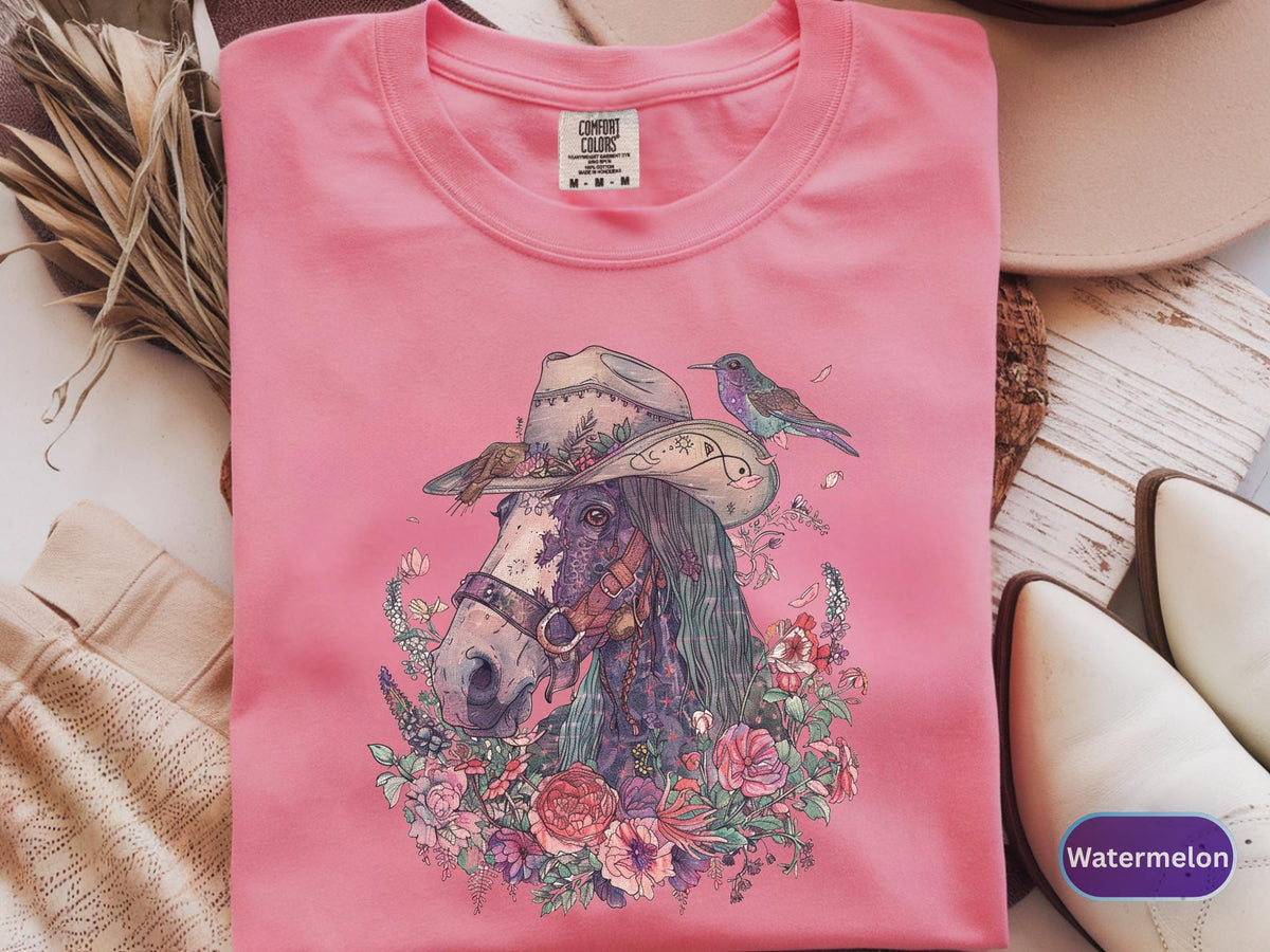 May include: Pink t-shirt with a detailed illustration of a horse wearing a cowboy hat, surrounded by flowers and a hummingbird. The shirt has the text "Comfort Colors" on the tag. The shirt is a size M-M-M. The shirt is a watermelon color.