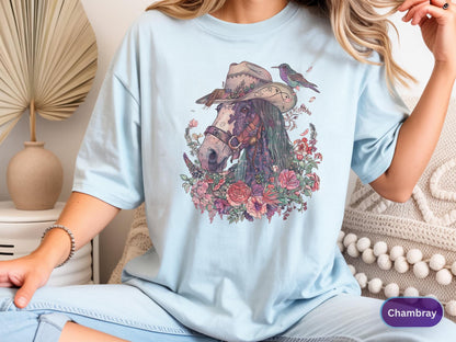 May include: Light blue t-shirt featuring a detailed illustration of a horse wearing a cowboy hat, surrounded by flowers and a small bird. The design is colorful and artistic, with a vintage aesthetic. The shirt is a relaxed fit.