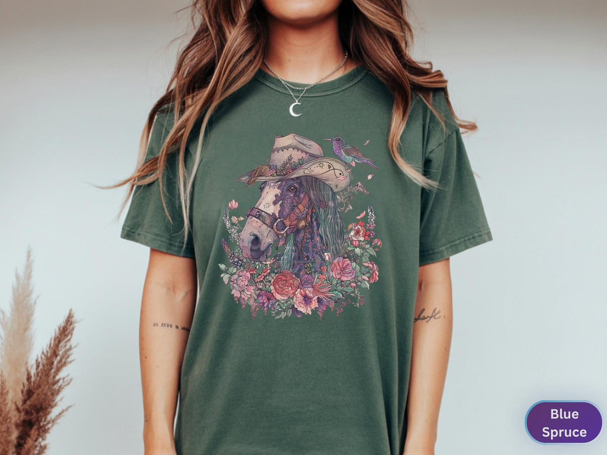 May include: A green t-shirt featuring a detailed illustration of a horse wearing a cowboy hat, surrounded by flowers and a hummingbird. The horse has a purple mane and is set against a backdrop of a floral arrangement. The shirt is labeled "Blue Spruce".