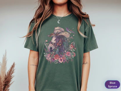 May include: A green t-shirt featuring a detailed illustration of a horse wearing a cowboy hat, surrounded by flowers and a hummingbird. The horse has a purple mane and is set against a backdrop of a floral arrangement. The shirt is labeled "Blue Spruce".
