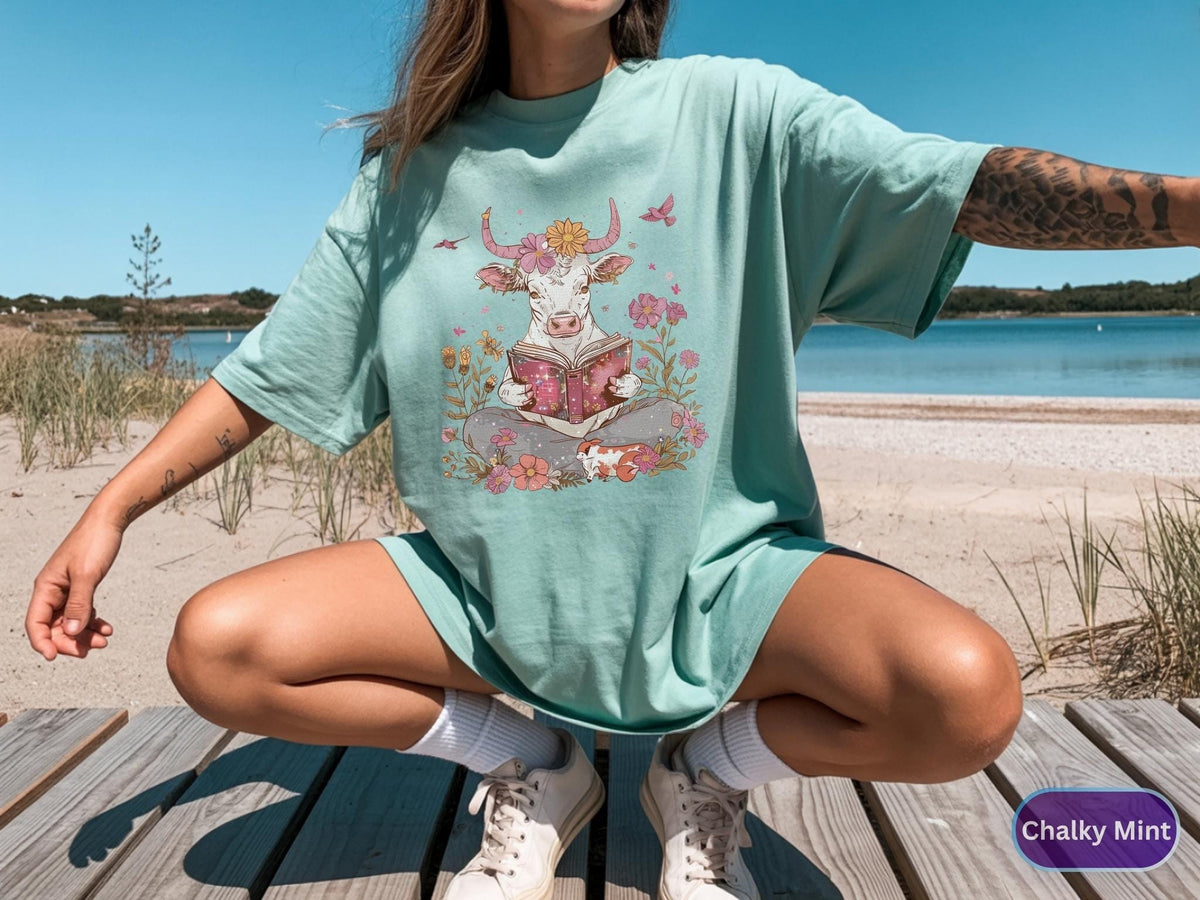 May include: A light teal t-shirt featuring a whimsical illustration of a white cow reading a book, surrounded by flowers and butterflies. The cow has a floral crown. The text "Chalky Mint" is in the lower right corner.