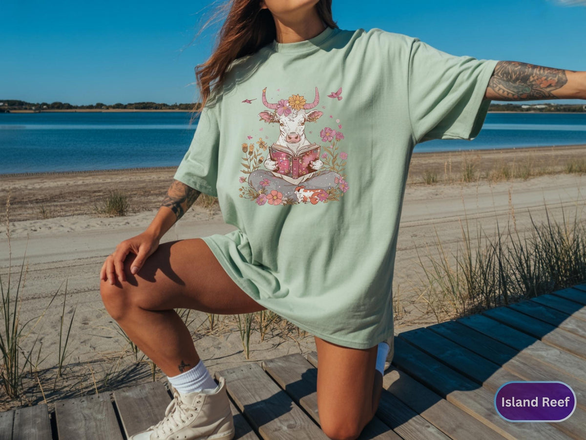 May include: A light green t-shirt featuring a whimsical illustration of a cow reading a book, surrounded by flowers and butterflies. The cow has a flower crown and is sitting cross-legged. The shirt is worn by a person.
