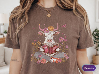 May include: Espresso-colored t-shirt featuring a whimsical illustration of a cow reading a book amidst flowers. The cow has floral accents and is surrounded by pink and yellow blooms. A small, patterned animal rests nearby. The shirt has a relaxed fit.