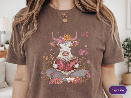 May include: Espresso-colored t-shirt featuring a whimsical illustration of a cow reading a book amidst flowers. The cow has floral accents and is surrounded by pink and yellow blooms. A small, patterned animal rests nearby. The shirt has a relaxed fit.