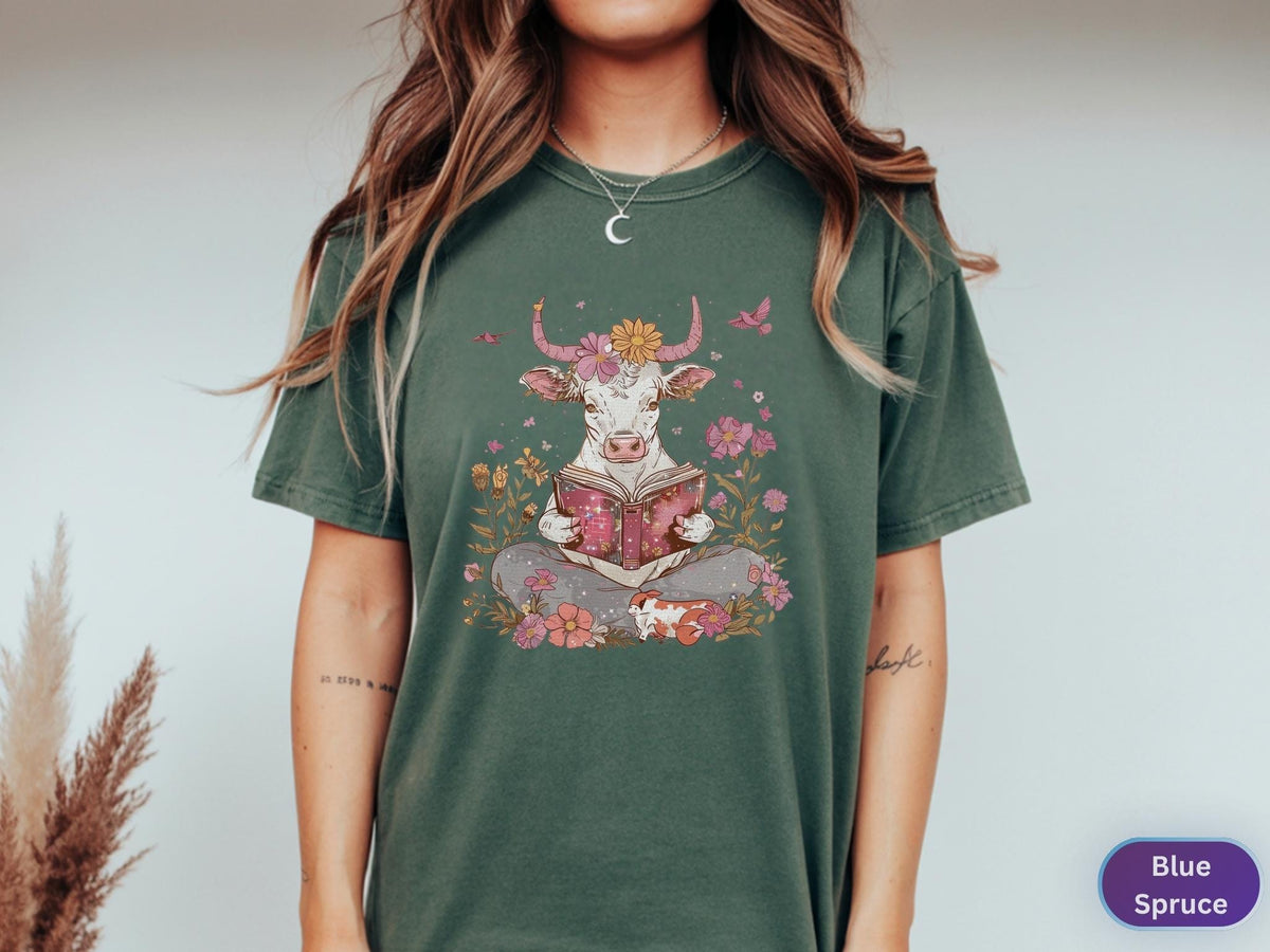 May include: A dark green t-shirt featuring a whimsical illustration of a cow reading a book, surrounded by flowers and butterflies. The cow has flower-adorned horns. The shirt is a comfortable, casual style.