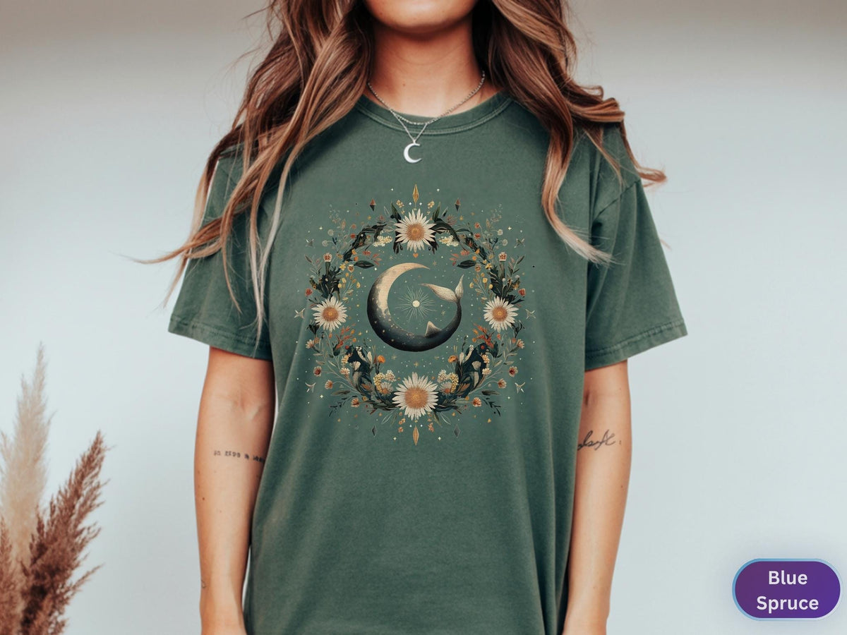 May include: A blue spruce t-shirt featuring a circular floral design with a crescent moon and a whale. The design includes sunflowers and other botanical elements. The shirt is a short-sleeved, crew neck style.