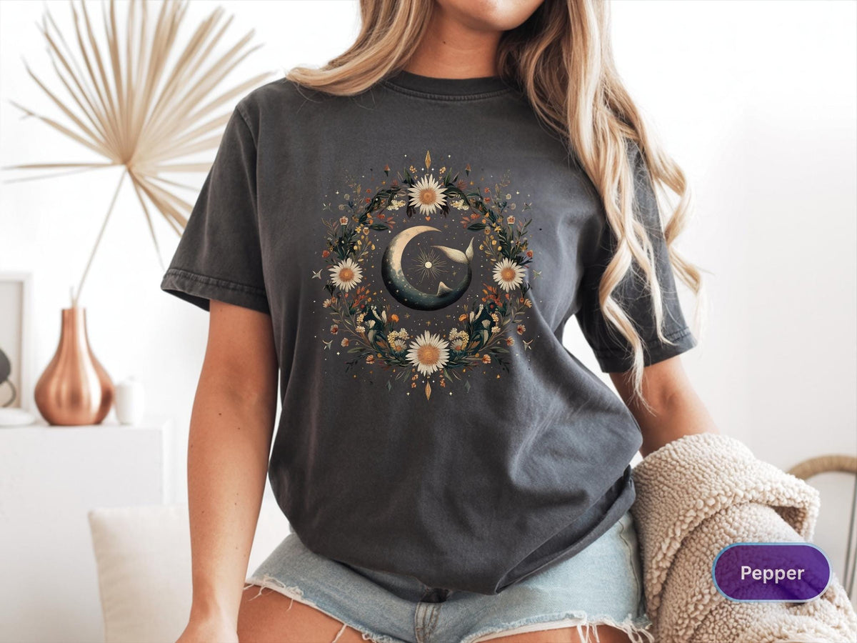 May include: Dark gray t-shirt featuring a celestial design. The graphic includes a crescent moon, a whale, and a floral wreath. The shirt is a relaxed fit and has short sleeves.