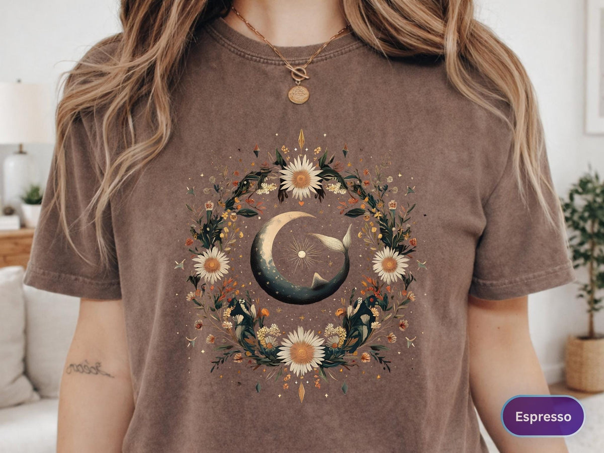 May include: Espresso-colored t-shirt featuring a celestial design. The graphic includes a crescent moon with a whale tail, surrounded by a floral wreath with daisies and other flowers. The shirt has a crew neck and short sleeves.