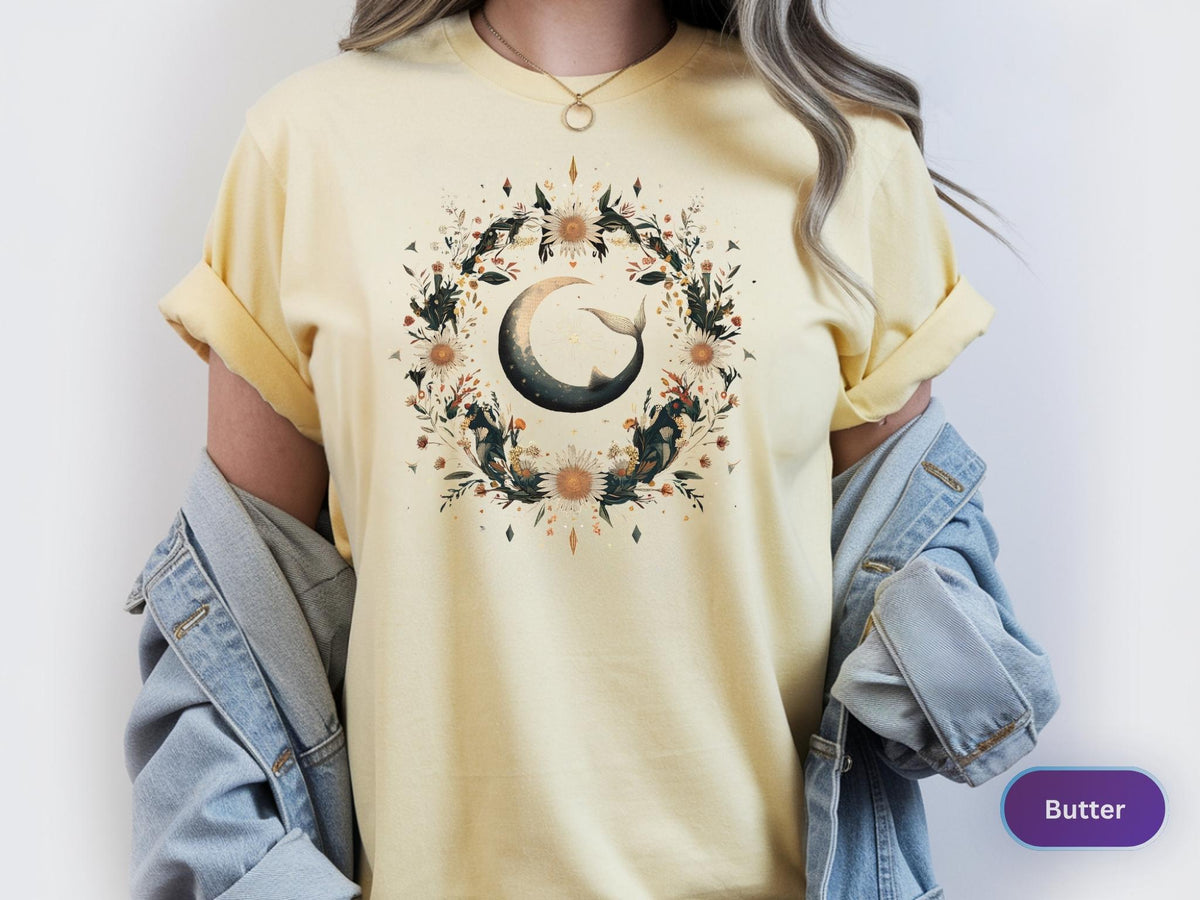 May include: Butter-colored t-shirt featuring a crescent moon design with a whale tail, surrounded by a floral wreath. The design includes dark green leaves, sunflowers, and other floral elements. The shirt has short sleeves and a relaxed fit.