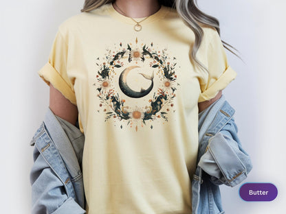 May include: Butter-colored t-shirt featuring a crescent moon design with a whale tail, surrounded by a floral wreath. The design includes dark green leaves, sunflowers, and other floral elements. The shirt has short sleeves and a relaxed fit.