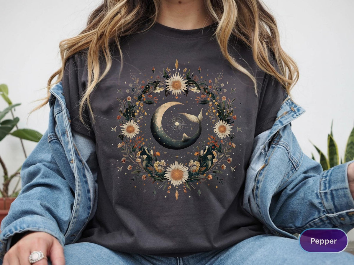 May include: Dark gray t-shirt featuring a celestial design. A crescent moon and whale are surrounded by a floral wreath of daisies and foliage. The word "Pepper" is visible on the bottom right.