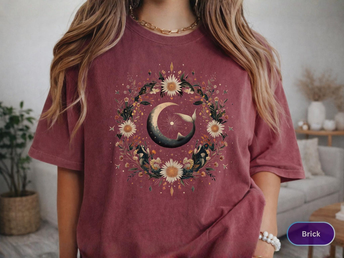 May include: Brick-colored t-shirt featuring a floral wreath design with a crescent moon and sun. The wreath includes sunflowers and other flowers. The shirt has short sleeves and a relaxed fit.