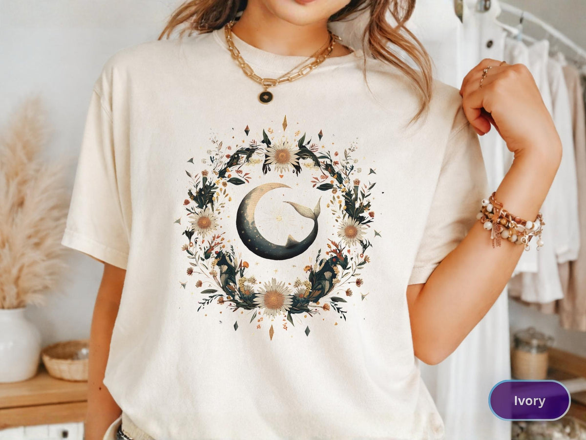 May include: Ivory-colored t-shirt featuring a watercolor design of a crescent moon with a whale tail, surrounded by a floral wreath. The design includes sunflowers, leaves, and other botanical elements. The shirt is a relaxed fit.