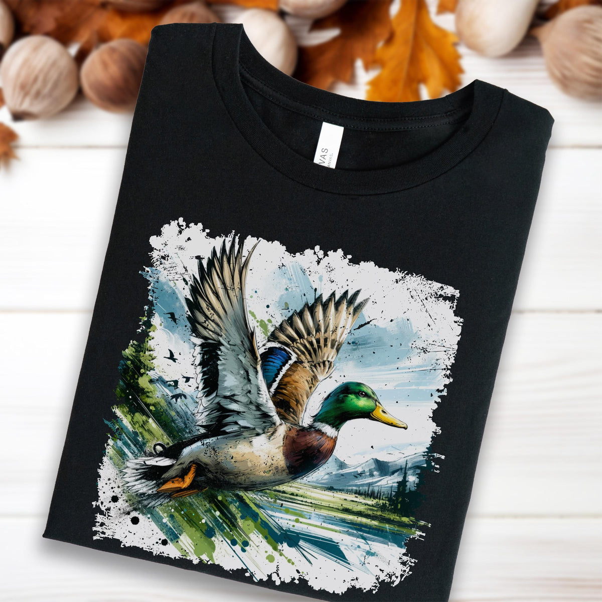 Black unisex tee featuring a bold watercolor illustration of a mallard duck in mid-flight with wings spread wide, set against a splattered paint background of mountains, trees, and a lake. Vivid green head, blue wing patch, and rust chest detail. Perfect duck hunting gift or wildlife nature shirt for outdoors lovers.
