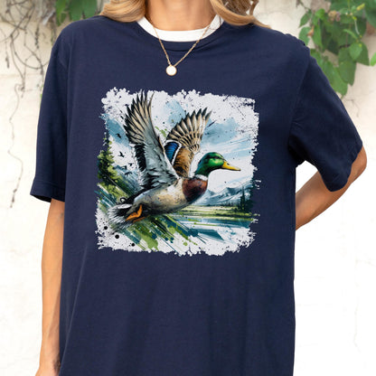 May include: Navy blue t-shirt featuring a detailed illustration of a mallard duck in flight. The duck has green, blue, and brown feathers, with wings spread wide. The artwork is set against a backdrop of a landscape with trees and mountains.