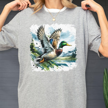 May include: A light gray t-shirt featuring a watercolor painting of a mallard duck in flight. The artwork includes a green-headed duck with outstretched wings, set against a backdrop of a lake, trees, and mountains.