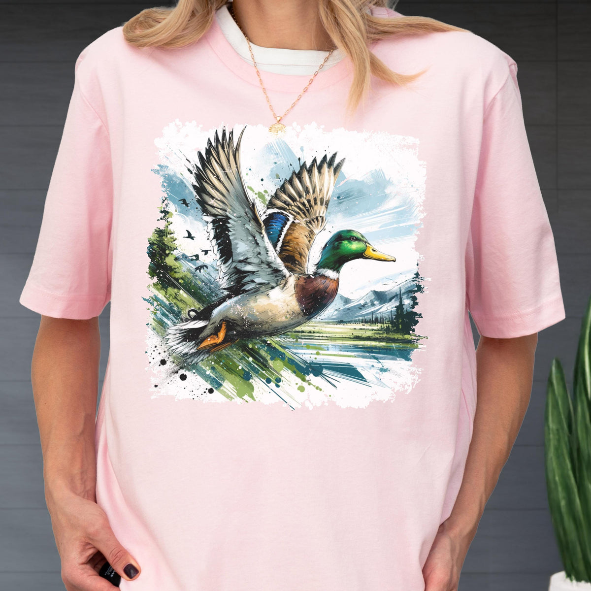 May include: Pink t-shirt featuring a watercolor illustration of a mallard duck in flight. The duck has green, brown, and blue feathers, with a yellow beak. The background includes a landscape with trees, water, and mountains.