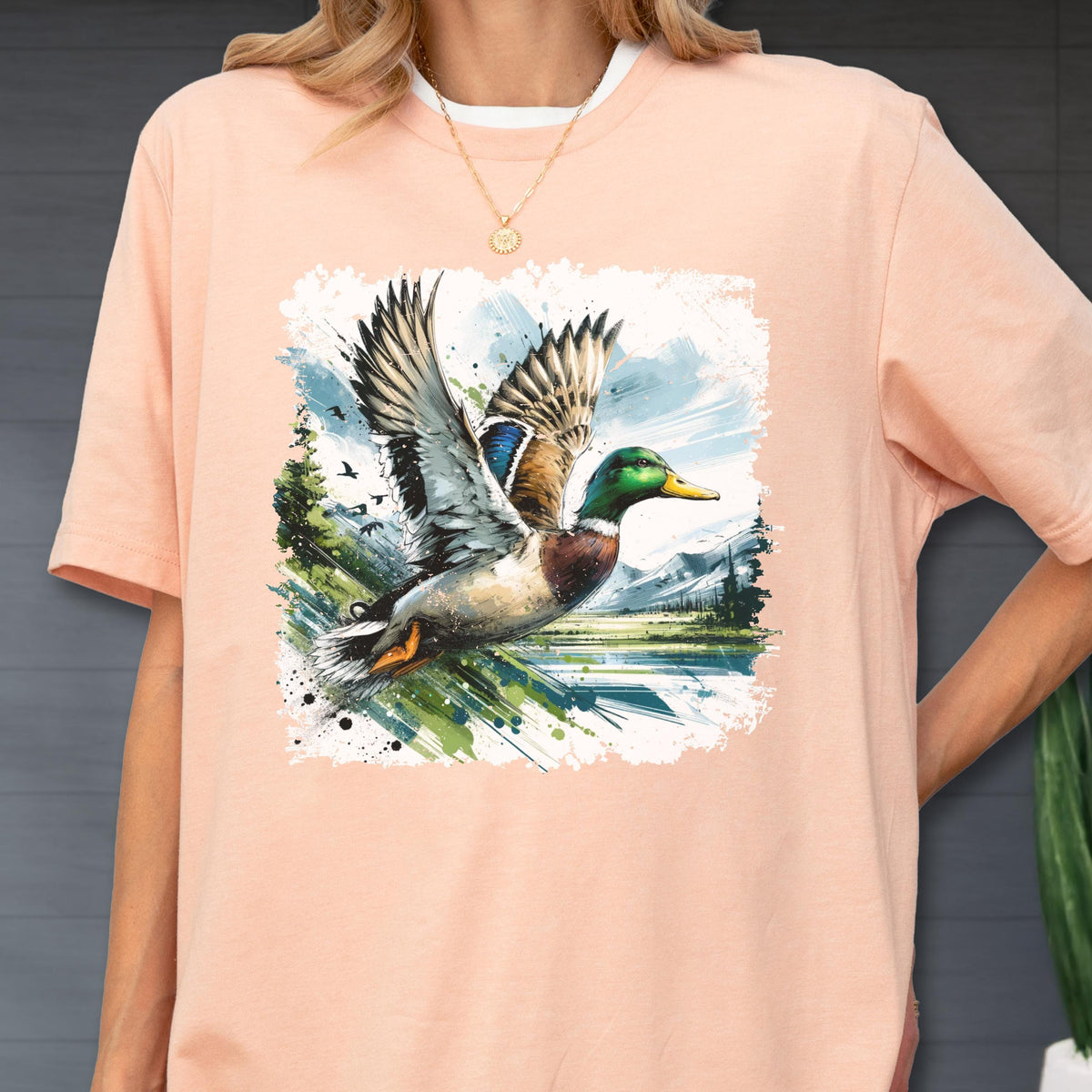 May include: A peach-colored t-shirt featuring a watercolor illustration of a mallard duck in flight. The duck has green, brown, and blue feathers, with wings spread wide. The background includes a lake, mountains, and trees.