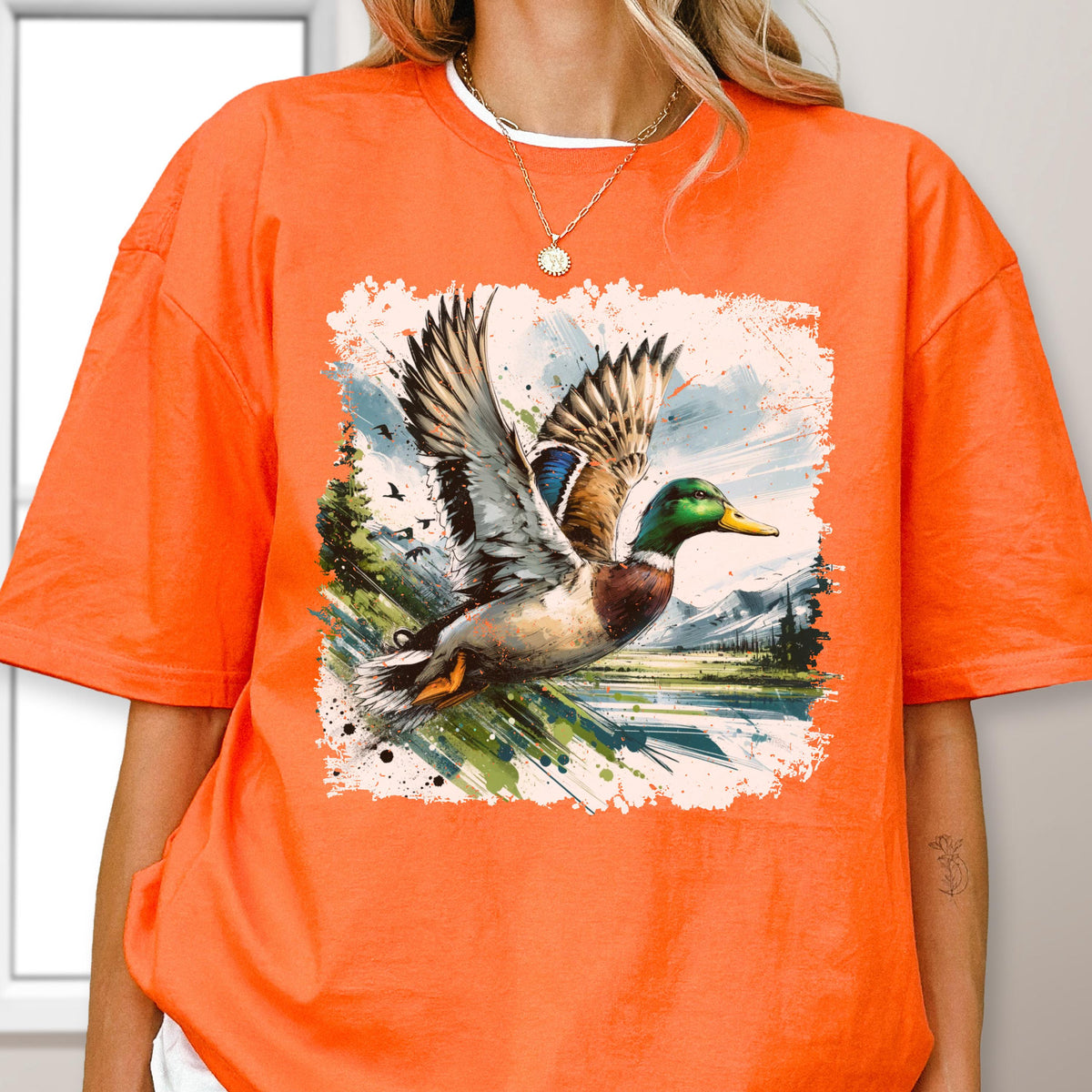 May include: Orange t-shirt featuring a watercolor illustration of a mallard duck in flight. The duck has green, brown, and blue feathers, with a yellow beak. The background includes a lake, trees, and mountains.