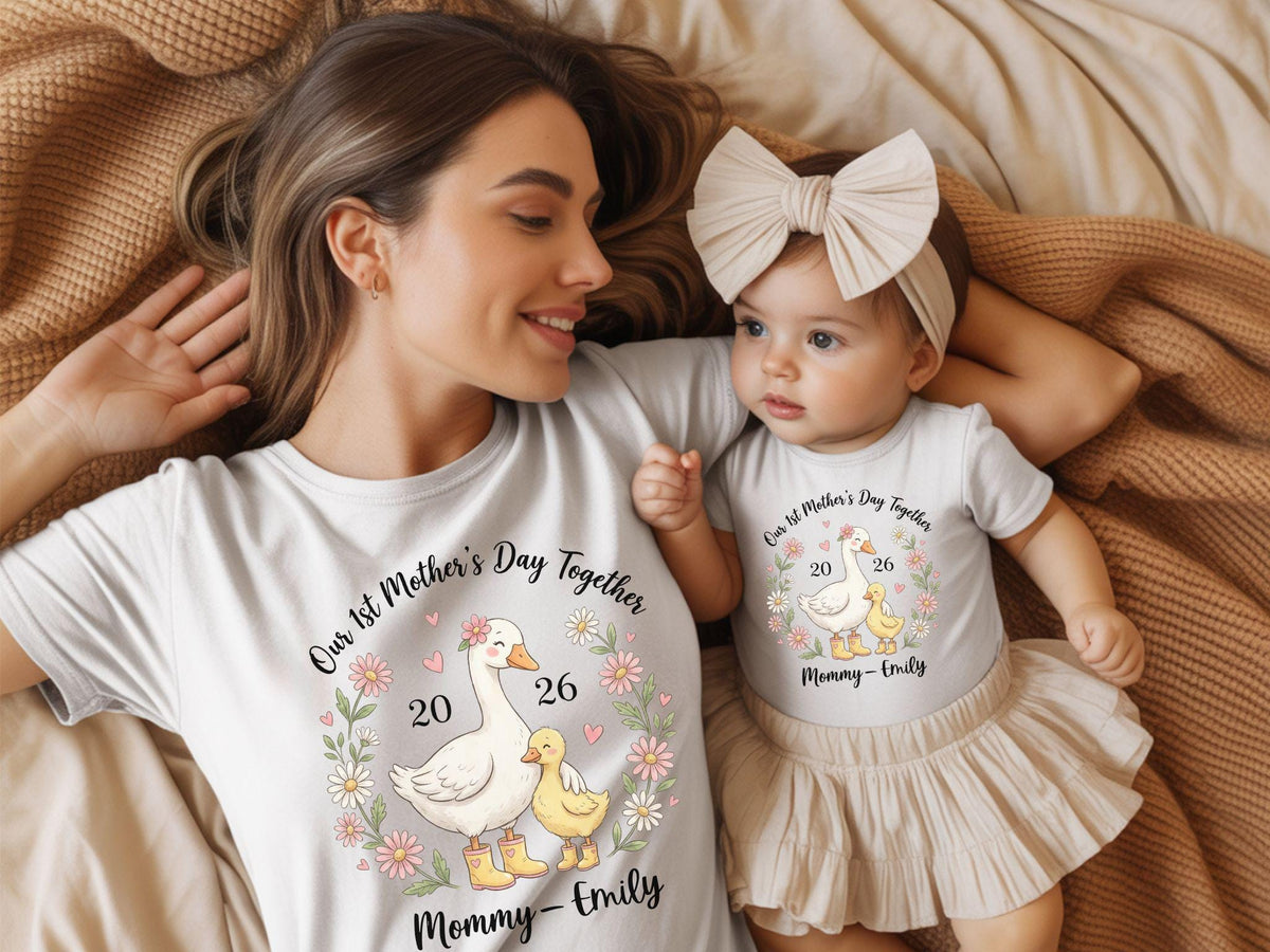 May include: Matching white t-shirts and onesie with a floral goose design. The shirts read "Our 1st Mother's Day Together" with the names "Mommy - Emily" and the numbers "20 26". The baby wears a beige skirt and headband.