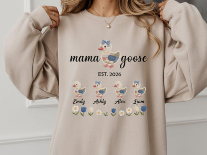 May include: Beige sweatshirt with a Mama Goose design. The design features a goose with a blue bow, the text "mama goose EST. 2026", and four ducklings with names. Floral and leaf accents are at the bottom.