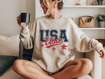 May include: A beige sweatshirt with the word "USA" in navy blue, distressed font, and "Usa" in red script. Two red stars are below the text. The person is holding a black mug.
